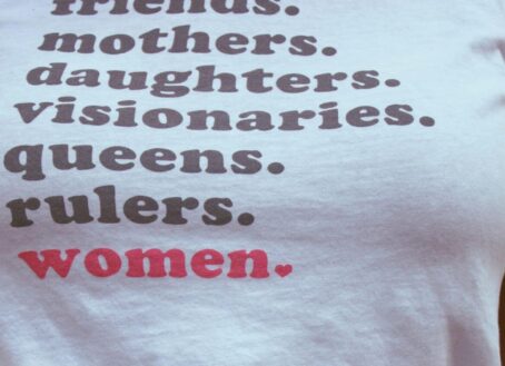 Young woman wearing a white t-shirt with friends, mothers, daughters, visionaries, queens, rulers, and women in bold print.